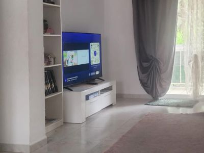 Vista parcial del salón con televisor en mueble blanco, estantería y acceso a terraza con cortinas grises y traslúcidas, sobre suelo de baldosas claras y alfombra.