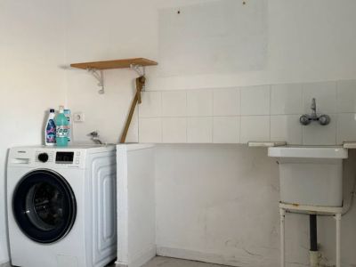 Zona de lavadero funcional con lavadora moderna, fregadero utilitario básico y salpicadero de azulejos blancos. Incluye suelo de baldosas claras y una sencilla estantería de madera.