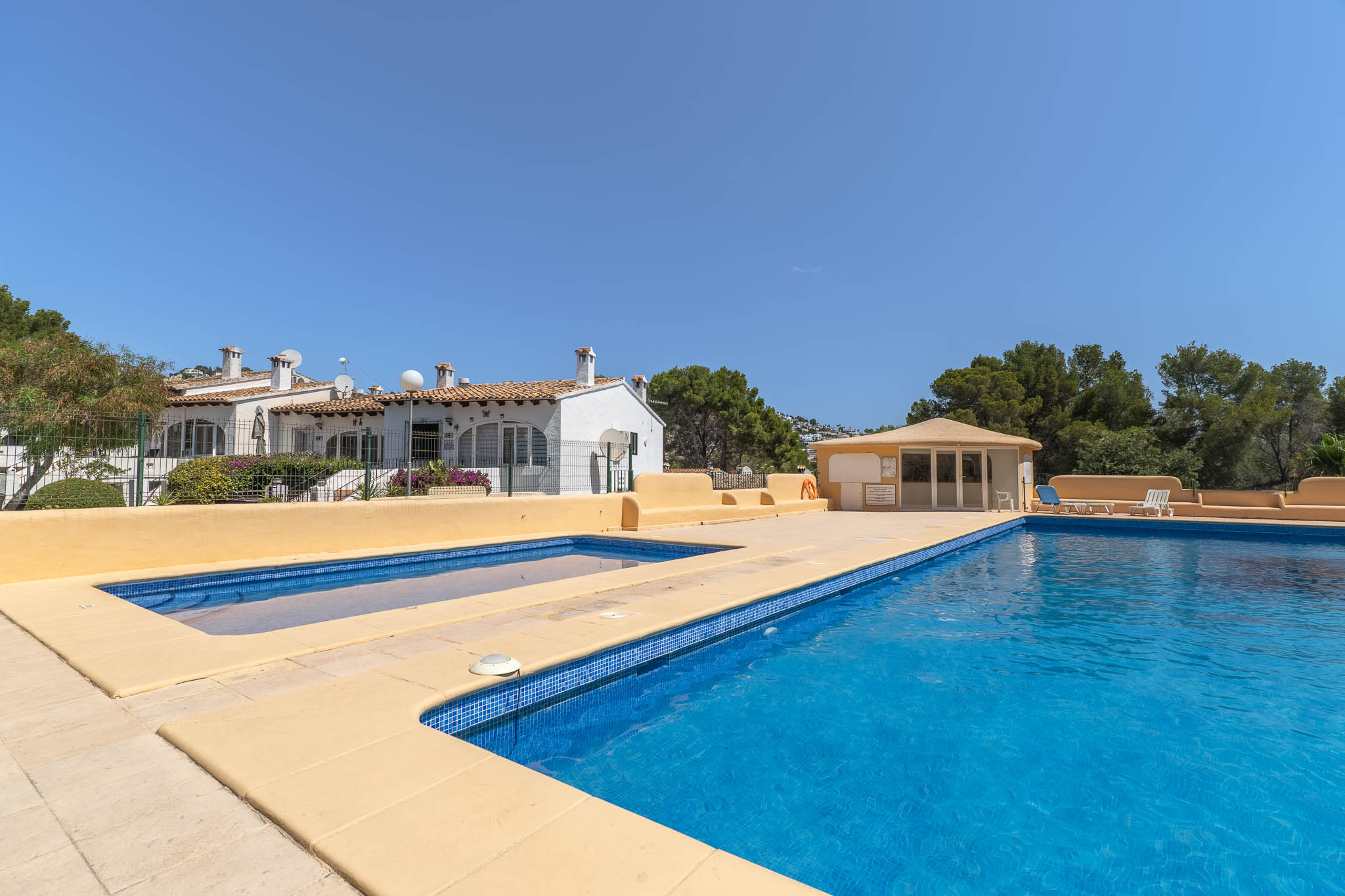 Amplia piscina comunitaria con mosaico azul y piscina infantil adyacente, rodeada de pavimento claro. Al fondo, casas mediterráneas y vegetación bajo un cielo despejado.