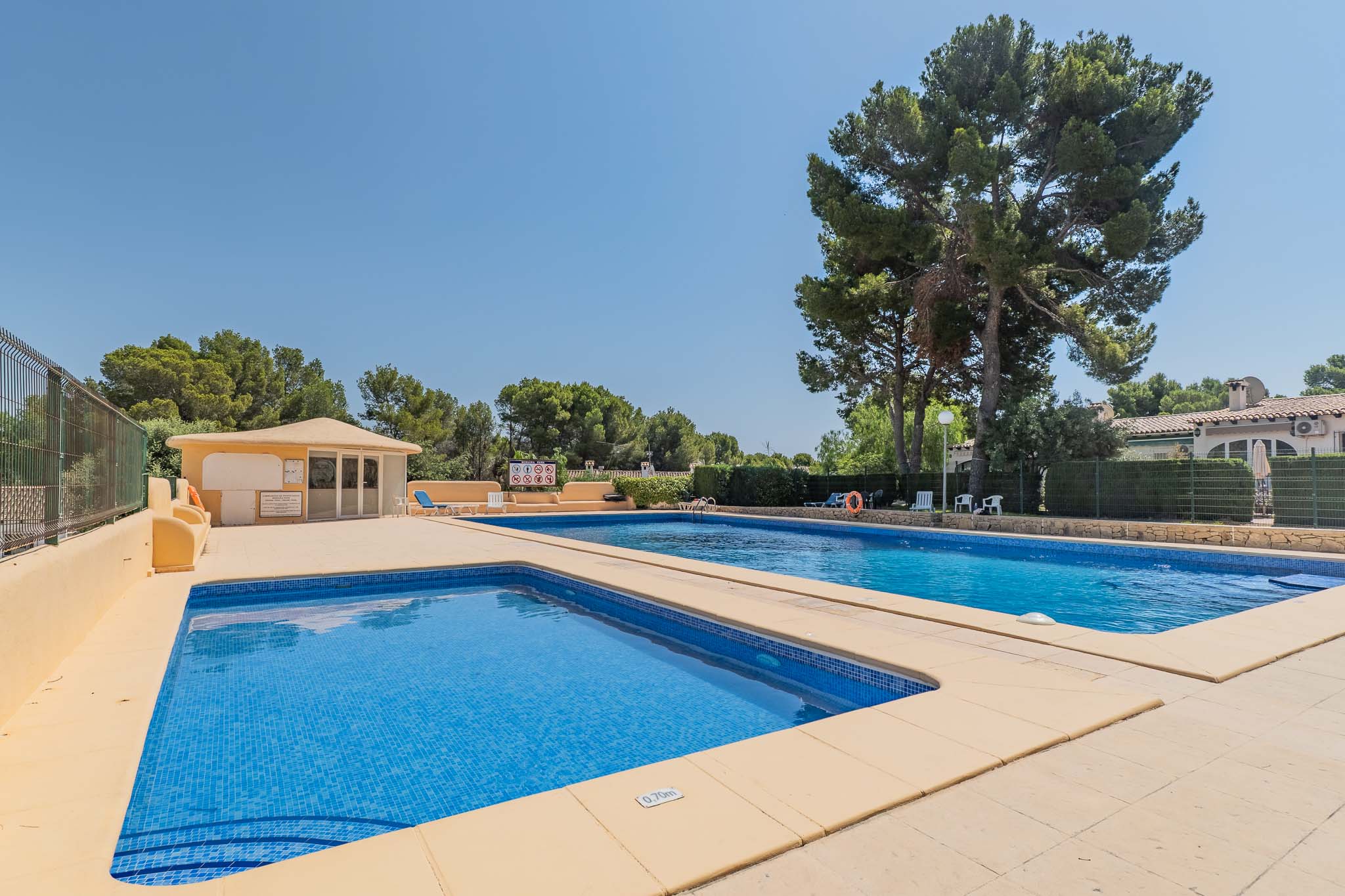 Amplia piscina comunitaria con un área infantil de 0.70m de profundidad, rodeada de frondosos pinos y un edificio de servicios, bajo un cielo despejado.