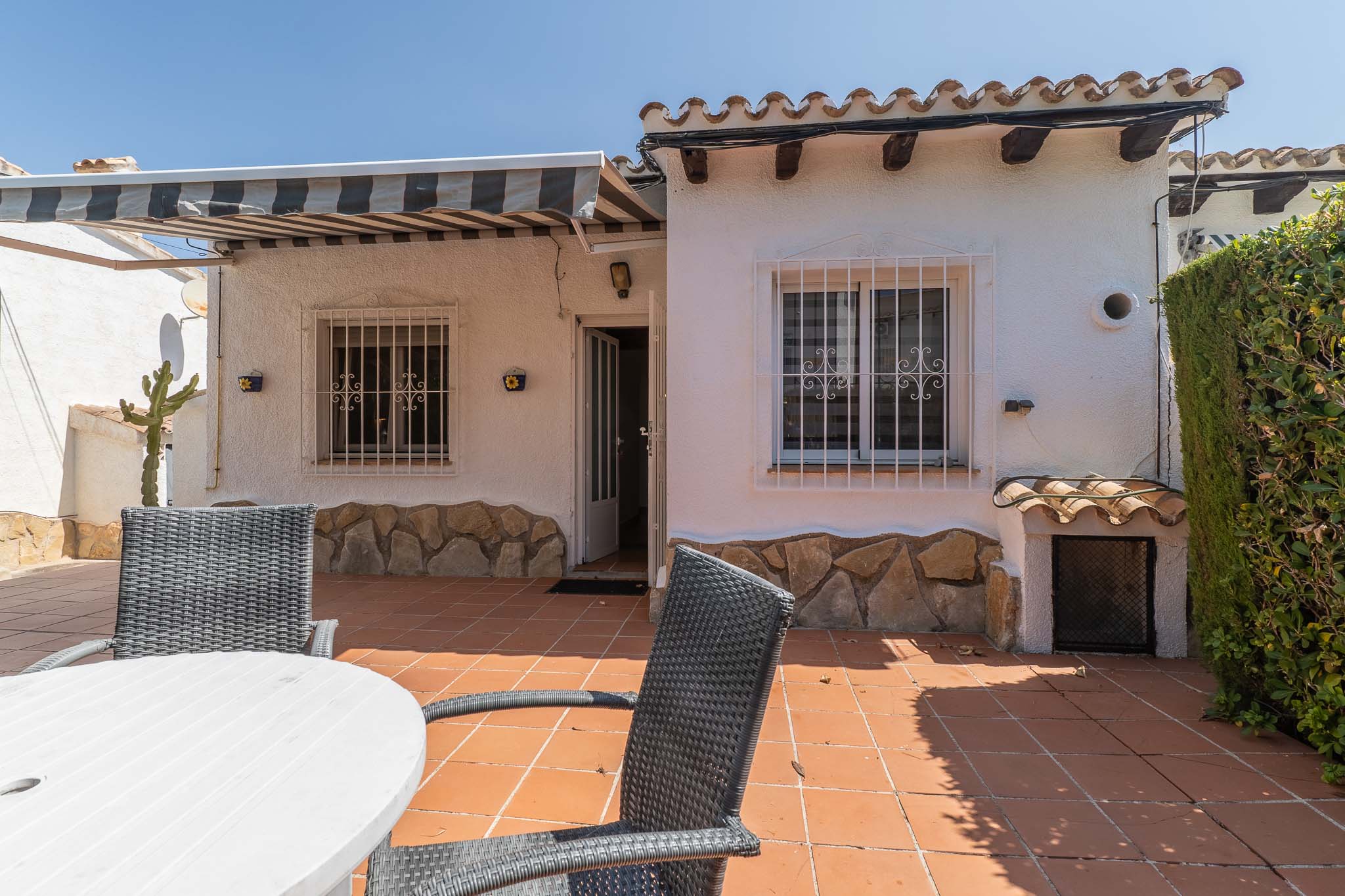 Amplia terraza exterior con suelo de baldosas de terracota, mobiliario de comedor y toldo retráctil. Fachada blanca con zócalo de piedra, ventanas con rejas y tejas tradicionales.