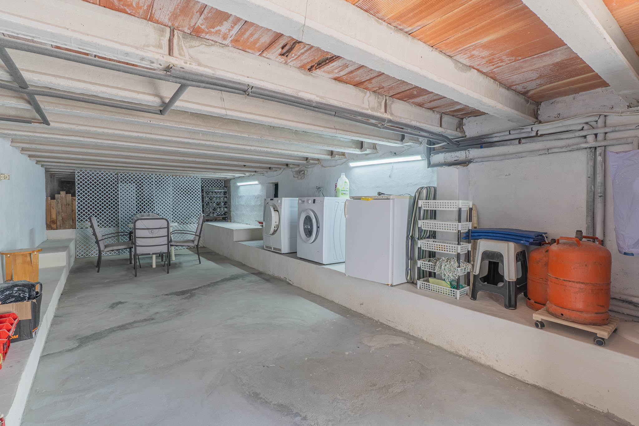 Amplio espacio inferior con zona de lavandería equipada, almacenamiento y un área de estar informal. Destaca su techo con vigas de hormigón y bovedillas cerámicas expuestas.