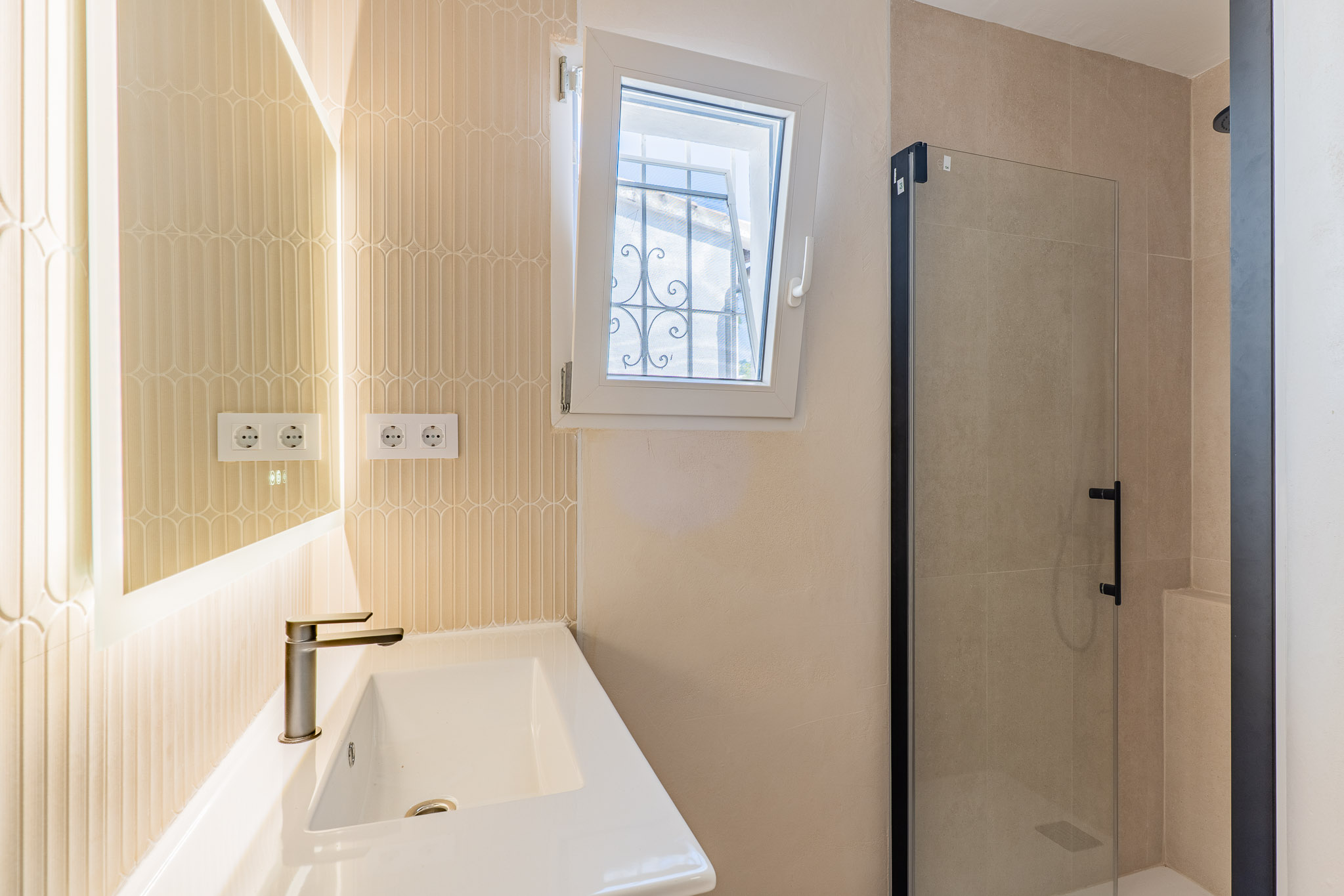 Baño moderno con lavabo cerámico, grifo bronce y espejo retroiluminado. Destaca la pared de azulejos verticales, ventana abatible con reja exterior y ducha con mampara de cristal y perfil negro.