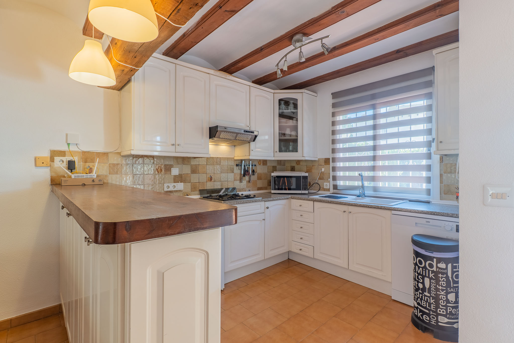 Cocina funcional con mobiliario blanco y encimera de madera. Destacan las vigas vistas en el techo, el suelo de terracota y la ventana con estor de lamas.