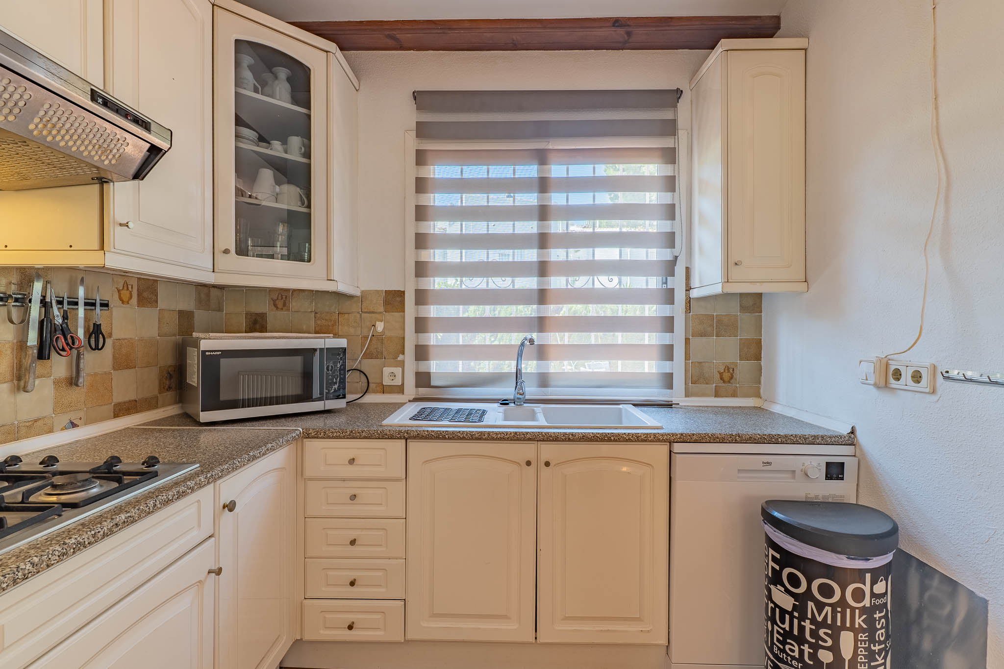 Cocina funcional con muebles de color crema, encimera de granito y salpicadero de azulejos. Incluye fregadero doble bajo ventana con persiana y electrodomésticos integrados.