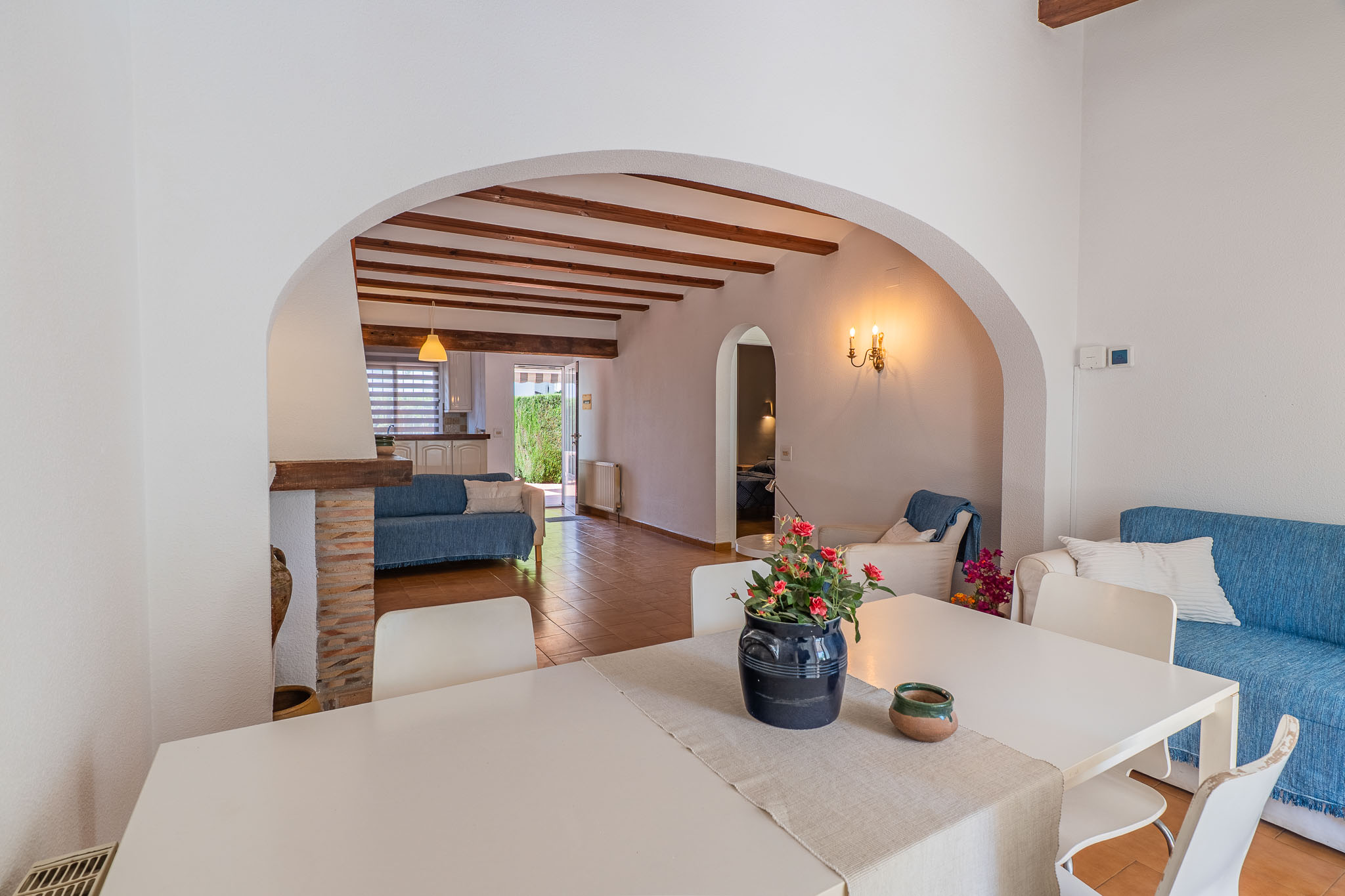 Comedor con mesa blanca, conectado por un arco a un salón con vigas de madera vistas, suelo de terracota y sofá azul, con acceso a la cocina y al jardín.