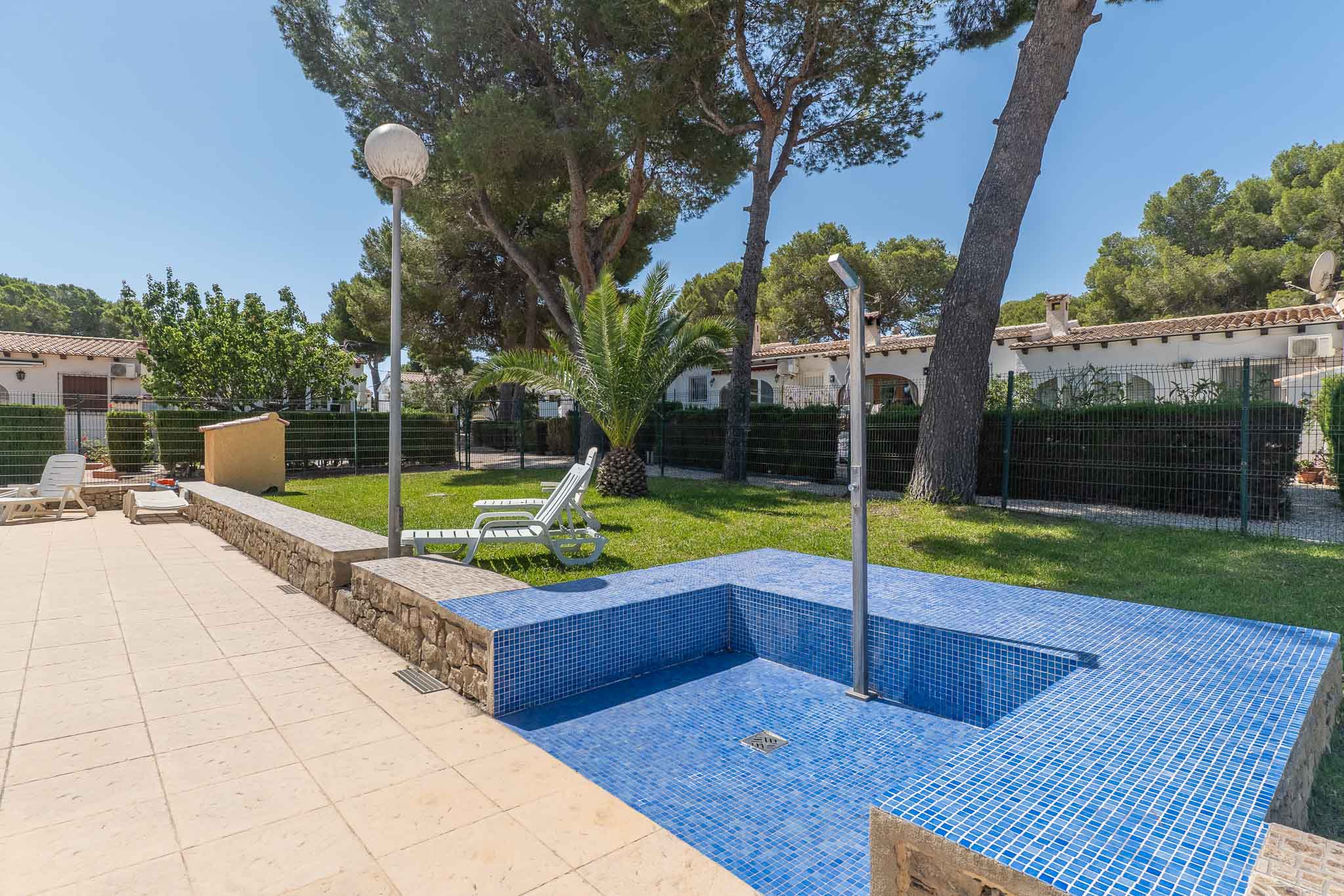 Detalle de la zona de piscina comunitaria con azulejos de mosaico azul, ducha exterior y pavimento claro, enmarcada por césped y vegetación.