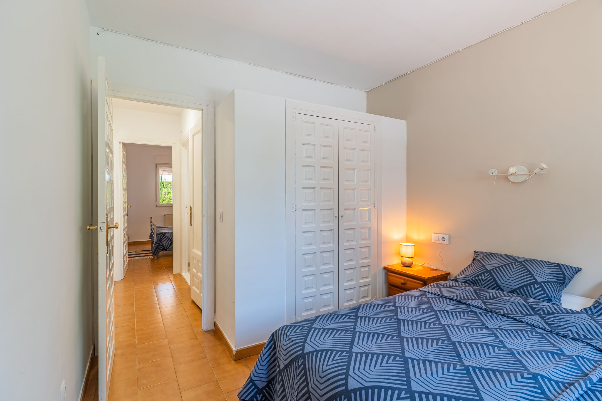 Dormitorio con cama de patrón geométrico azul, mesita de noche de madera con lámpara y armario empotrado blanco con paneles. Un pasillo con suelo de terracota conduce a una estancia adyacente.