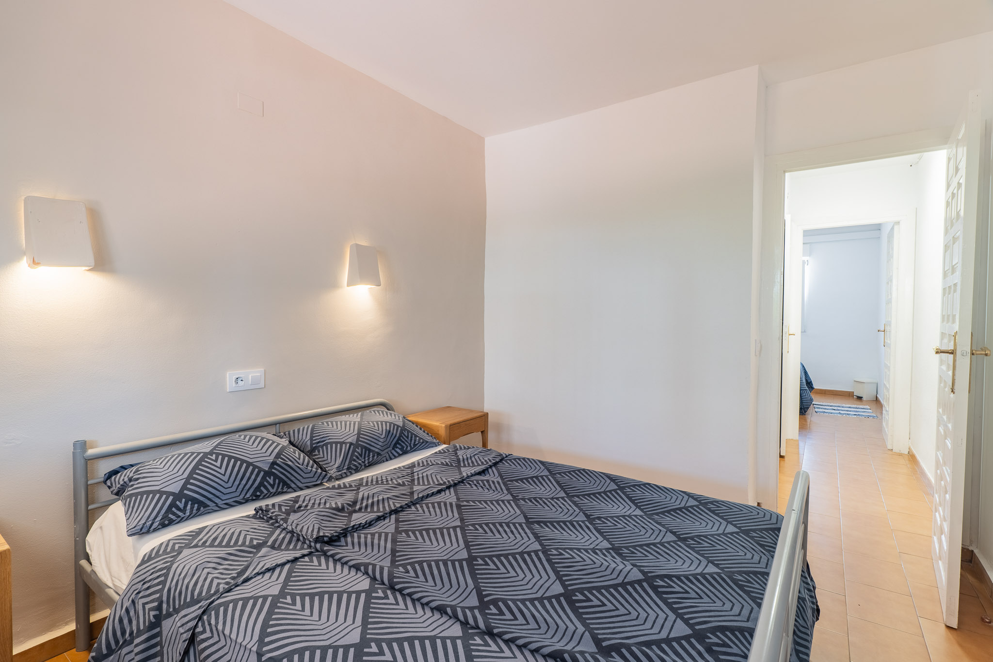 Dormitorio con cama doble de estructura metálica gris y ropa de cama estampada. Dos apliques de pared iluminan sobre una mesita de noche de madera. Una puerta abierta muestra un pasillo con azulejos.