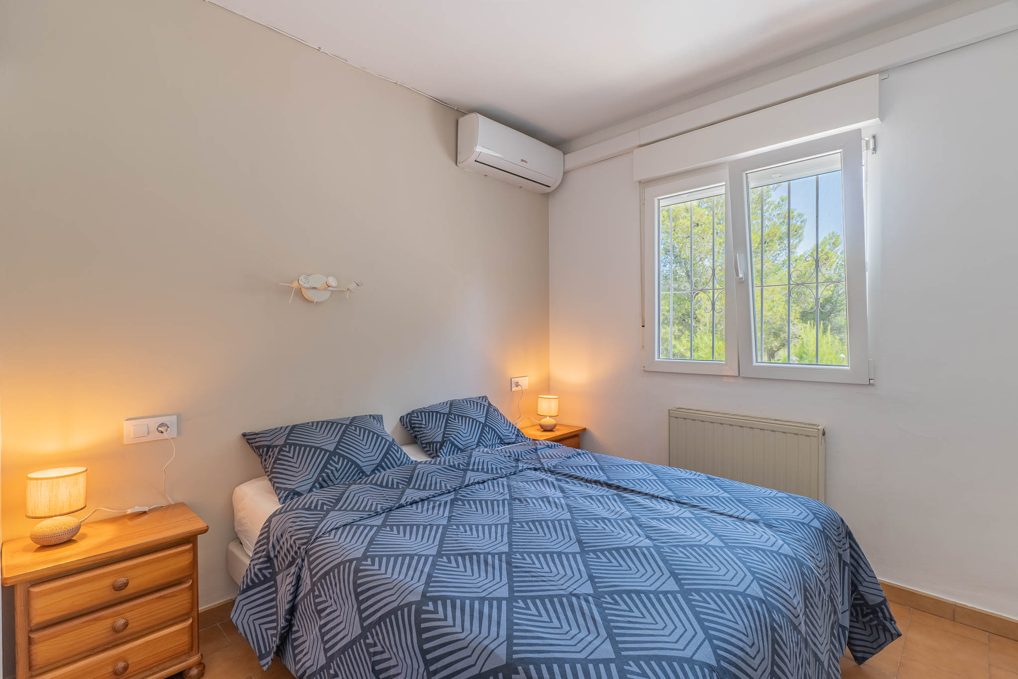 Dormitorio con cama doble, mesitas de noche de madera y aire acondicionado. Ventana con rejas y vistas a la vegetación, además de radiador.