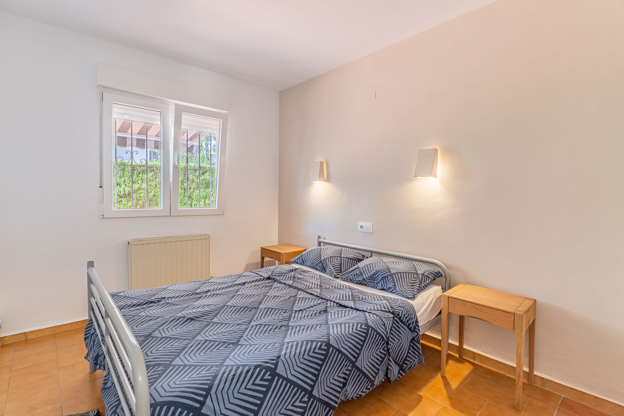 Dormitorio con cama doble, mesitas de noche de madera y suelo de terracota. La ventana con rejas ofrece vistas a la vegetación exterior y un radiador.