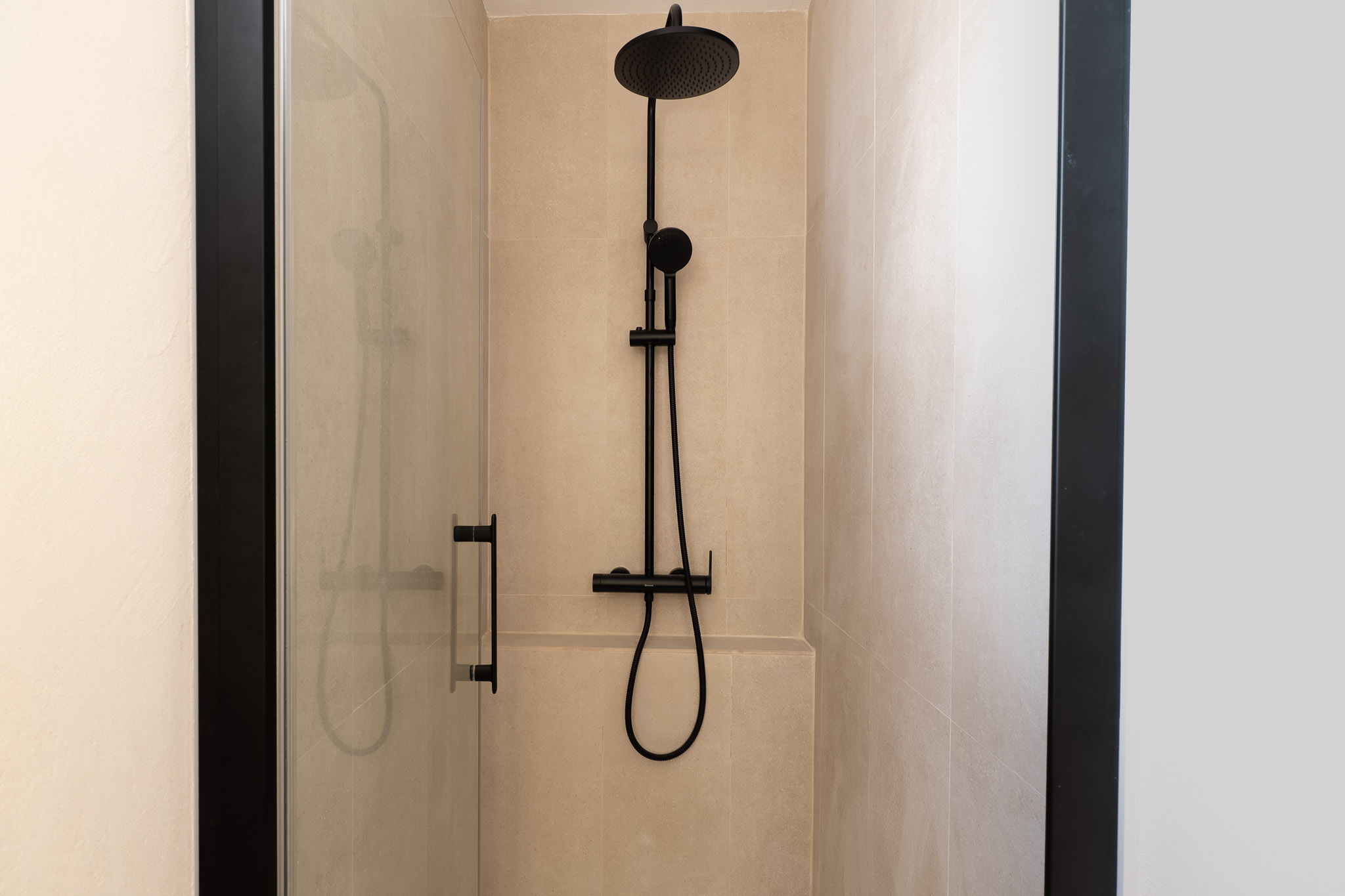 Ducha moderna con azulejos beige y grifería negra mate. La mampara de cristal transparente con perfilería oscura complementa el diseño contemporáneo del baño.