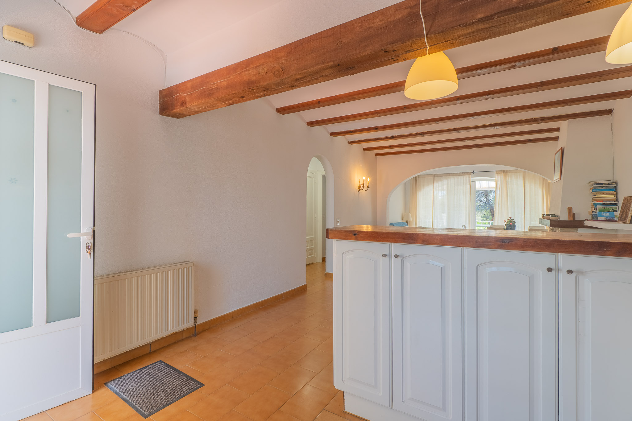 Entrada con puerta de cristal esmerilado y radiador, suelo de terracota, y techo con vigas de madera. Una barra americana con encimera de madera conecta con el luminoso salón.