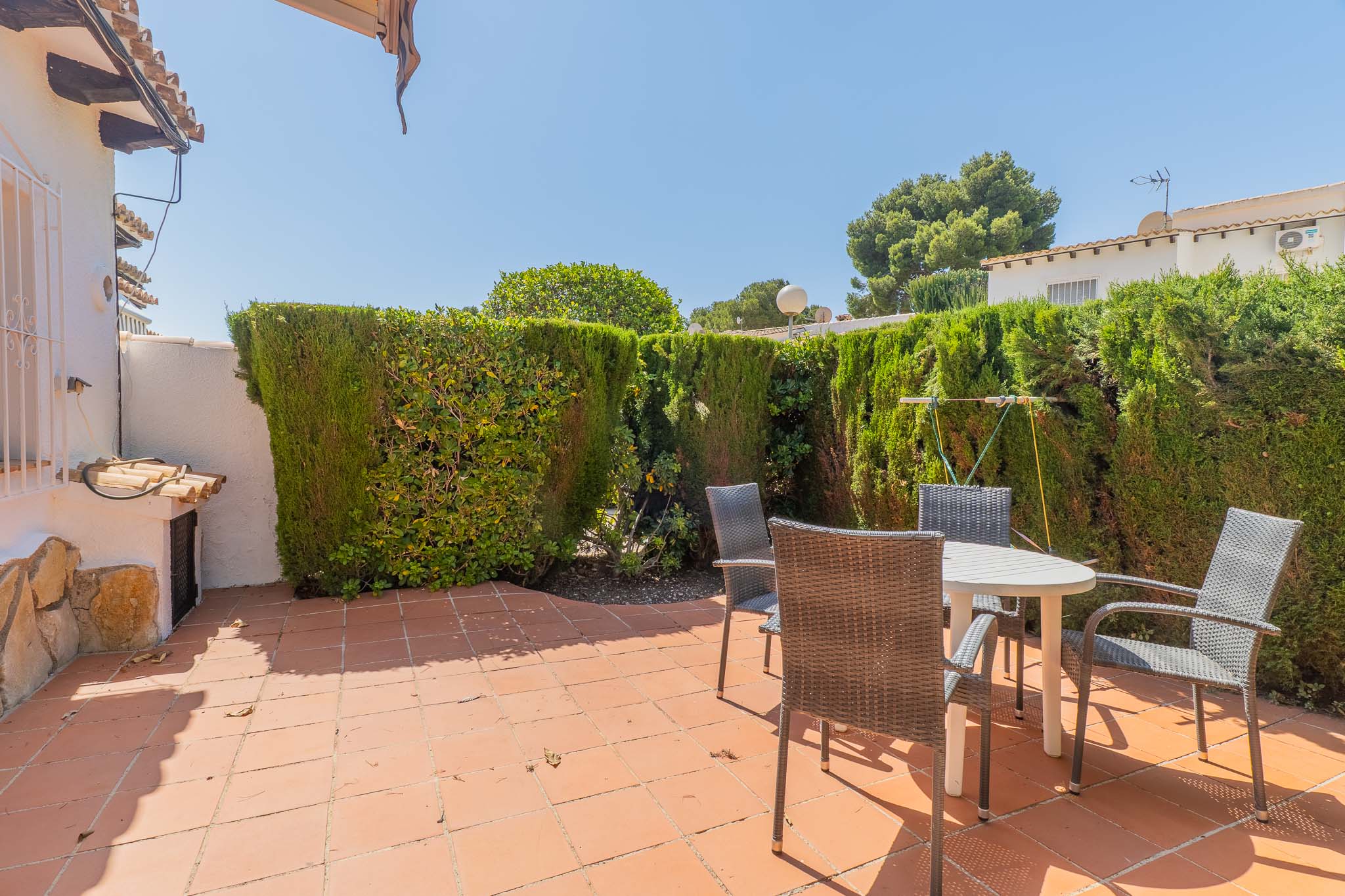 Patio trasero privado y soleado con suelo de baldosas de terracota, mesa redonda y sillas de jardín, rodeado por un seto alto que ofrece privacidad.