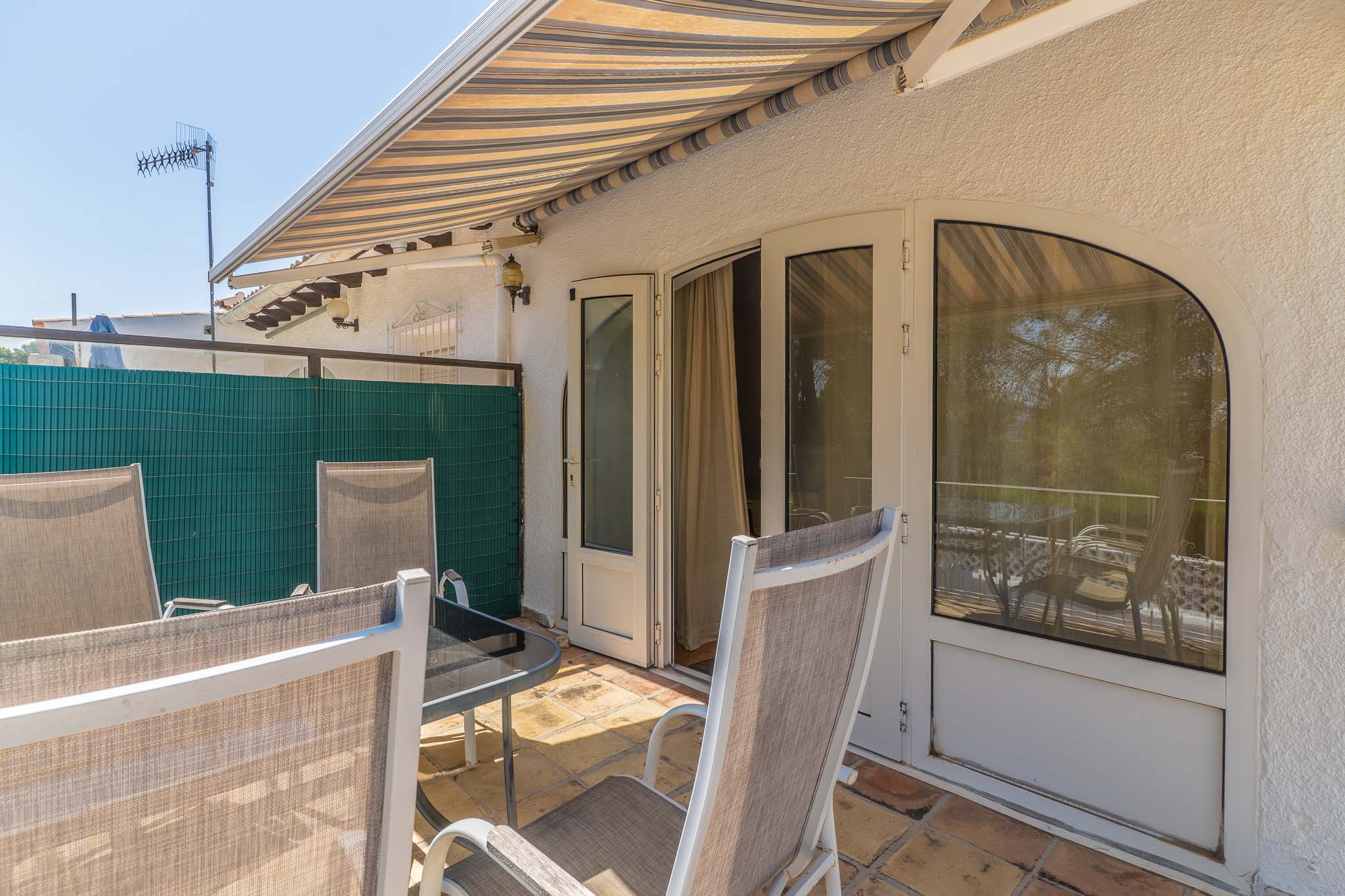 Soleada terraza exterior con suelo de terracota, toldo a rayas y mobiliario de jardín. Acceso directo al interior mediante puertas dobles y ventana arqueada.