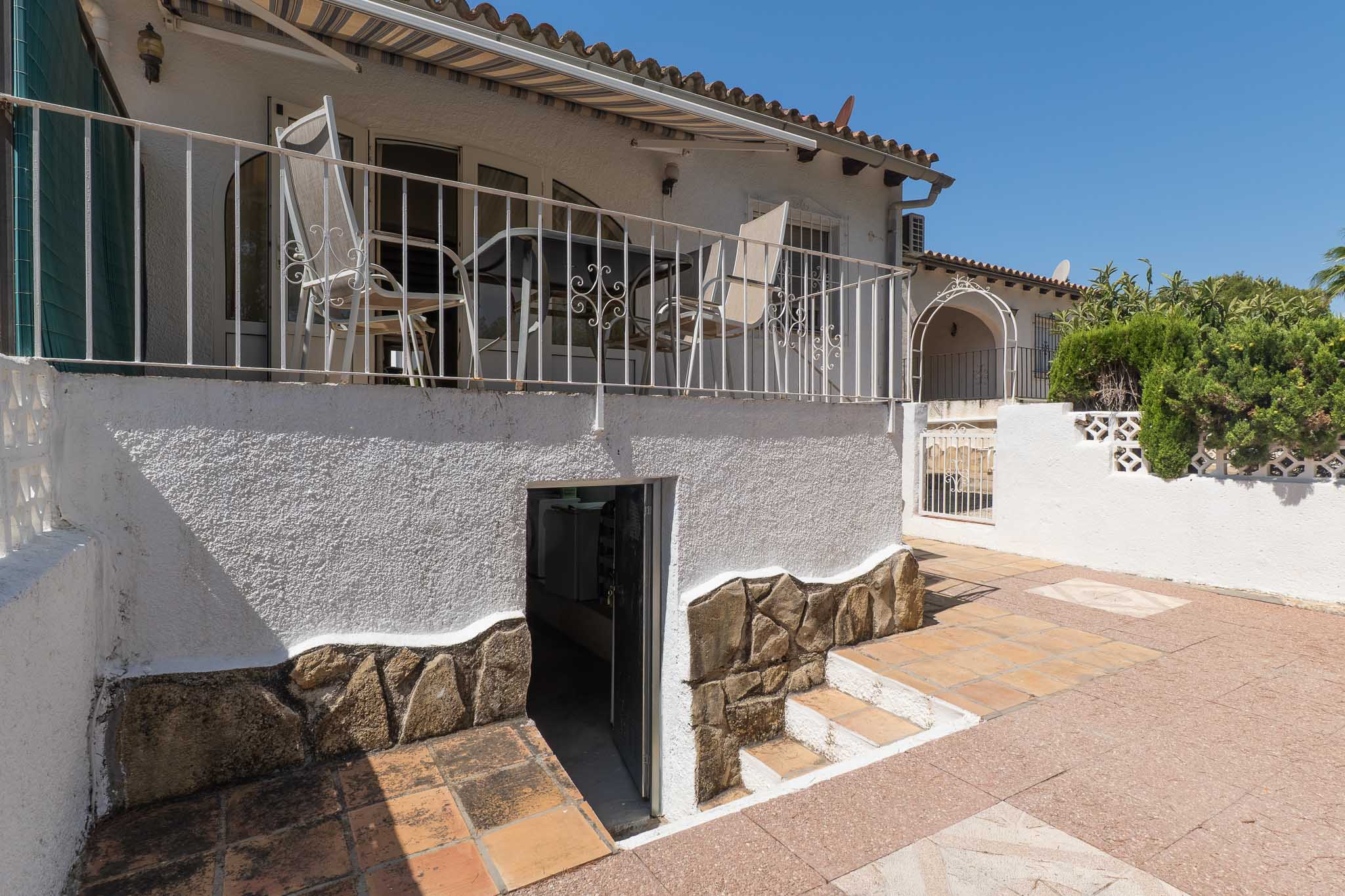 Terraza elevada con barandilla de hierro forjado y toldo, conectada a un patio inferior pavimentado con acceso a un trastero/lavandería. Muros con base de piedra decorativa.