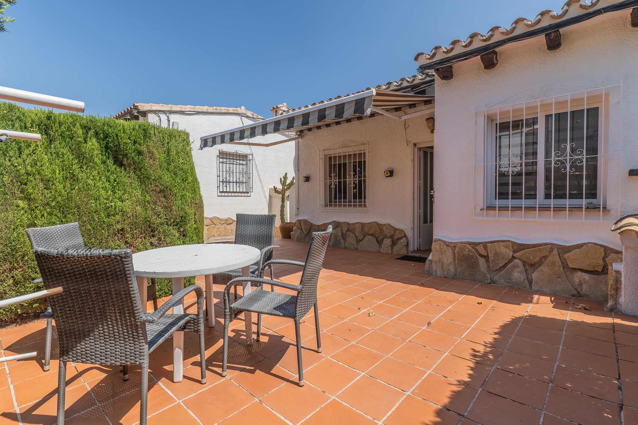Terraza exterior con suelo de baldosas de terracota, equipada con mesa blanca y sillas de mimbre gris. Un seto alto ofrece privacidad, mientras un toldo extensible sombrea la fachada con base de piedra.