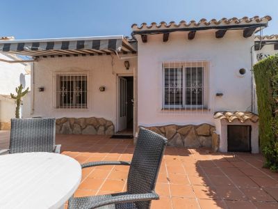 Amplia terraza exterior con suelo de baldosas de terracota, mobiliario de comedor y toldo retráctil. Fachada blanca con zócalo de piedra, ventanas con rejas y tejas tradicionales.