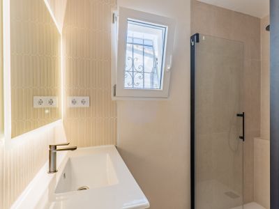 Baño moderno con lavabo cerámico, grifo bronce y espejo retroiluminado. Destaca la pared de azulejos verticales, ventana abatible con reja exterior y ducha con mampara de cristal y perfil negro.