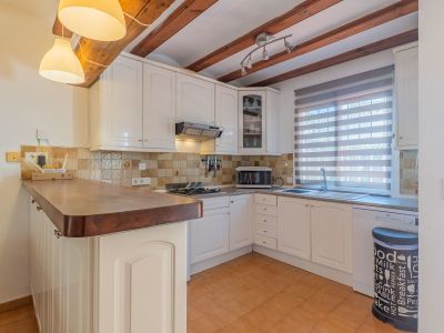 Cocina funcional con mobiliario blanco y encimera de madera. Destacan las vigas vistas en el techo, el suelo de terracota y la ventana con estor de lamas.