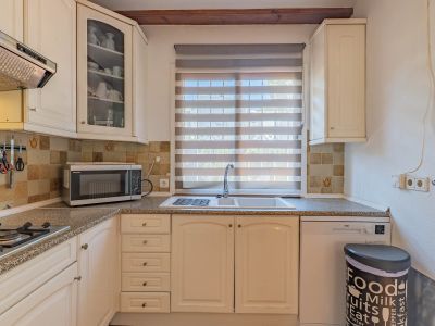 Cocina funcional con muebles de color crema, encimera de granito y salpicadero de azulejos. Incluye fregadero doble bajo ventana con persiana y electrodomésticos integrados.