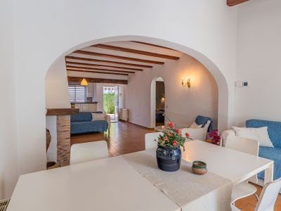 Comedor con mesa blanca, conectado por un arco a un salón con vigas de madera vistas, suelo de terracota y sofá azul, con acceso a la cocina y al jardín.