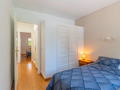 Dormitorio con cama de patrón geométrico azul, mesita de noche de madera con lámpara y armario empotrado blanco con paneles. Un pasillo con suelo de terracota conduce a una estancia adyacente.