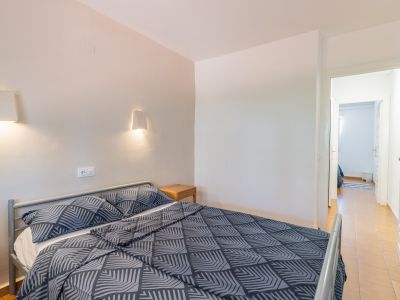 Dormitorio con cama doble de estructura metálica gris y ropa de cama estampada. Dos apliques de pared iluminan sobre una mesita de noche de madera. Una puerta abierta muestra un pasillo con azulejos.
