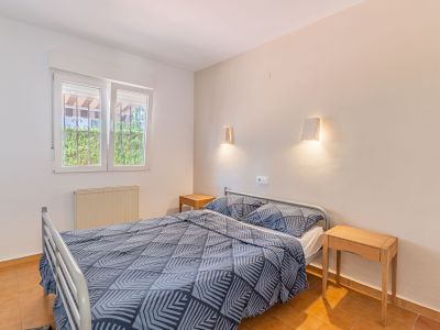 Dormitorio con cama doble, mesitas de noche de madera y suelo de terracota. La ventana con rejas ofrece vistas a la vegetación exterior y un radiador.