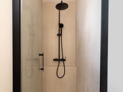 Ducha moderna con azulejos beige y grifería negra mate. La mampara de cristal transparente con perfilería oscura complementa el diseño contemporáneo del baño.