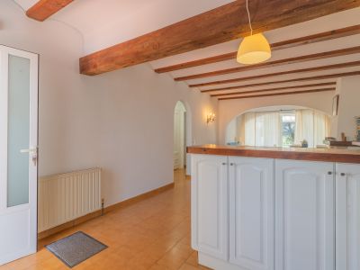 Entrada con puerta de cristal esmerilado y radiador, suelo de terracota, y techo con vigas de madera. Una barra americana con encimera de madera conecta con el luminoso salón.