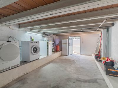 Espacio inferior funcional con lavadora, secadora y neveras, techo de vigas vistas y suelo de hormigón. Incluye salida exterior y zona de almacenaje.
