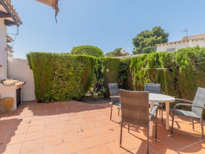 Patio trasero privado y soleado con suelo de baldosas de terracota, mesa redonda y sillas de jardín, rodeado por un seto alto que ofrece privacidad.
