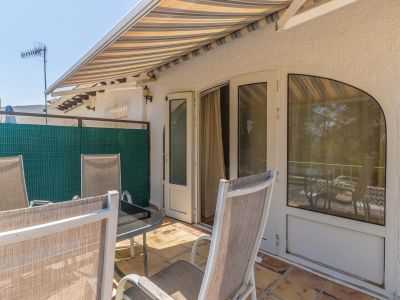 Soleada terraza exterior con suelo de terracota, toldo a rayas y mobiliario de jardín. Acceso directo al interior mediante puertas dobles y ventana arqueada.