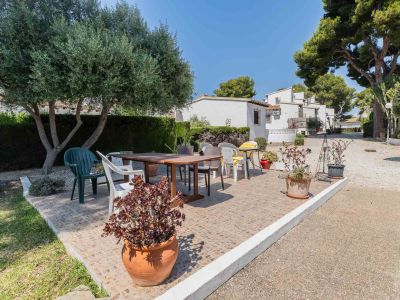 Soleado patio alicatado con mesa de comedor de madera y sillas variadas, flanqueado por un olivo y macetas. Edificios residenciales blancos se aprecian al fondo bajo un cielo despejado.