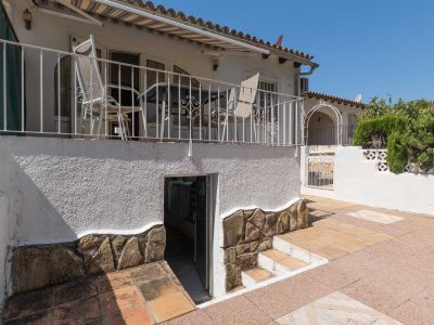 Terraza elevada con barandilla de hierro forjado y toldo, conectada a un patio inferior pavimentado con acceso a un trastero/lavandería. Muros con base de piedra decorativa.