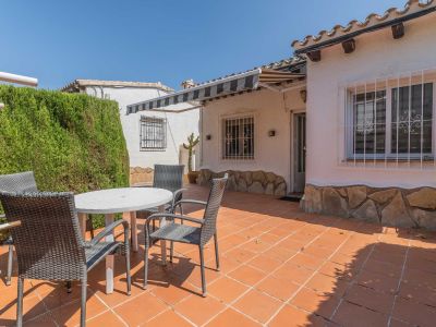 Terraza exterior con suelo de baldosas de terracota, equipada con mesa blanca y sillas de mimbre gris. Un seto alto ofrece privacidad, mientras un toldo extensible sombrea la fachada con base de piedra.