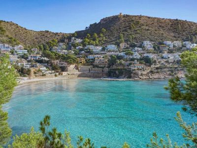 Vista panorámica de una bahía tranquila con aguas turquesas cristalinas, playa de arena y casas blancas en la ladera verde y arbolada, coronada por una torre de vigilancia histórica.