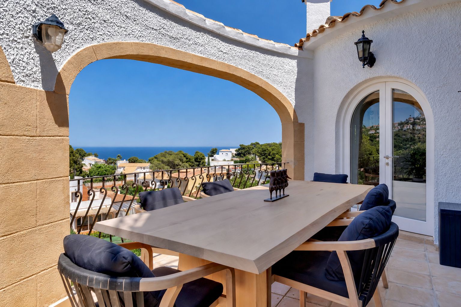 Amplia terraza comedor con mesa de madera clara y sillas con cojines azules. Un arco enmarca vistas panorámicas al mar Mediterráneo y la costa.