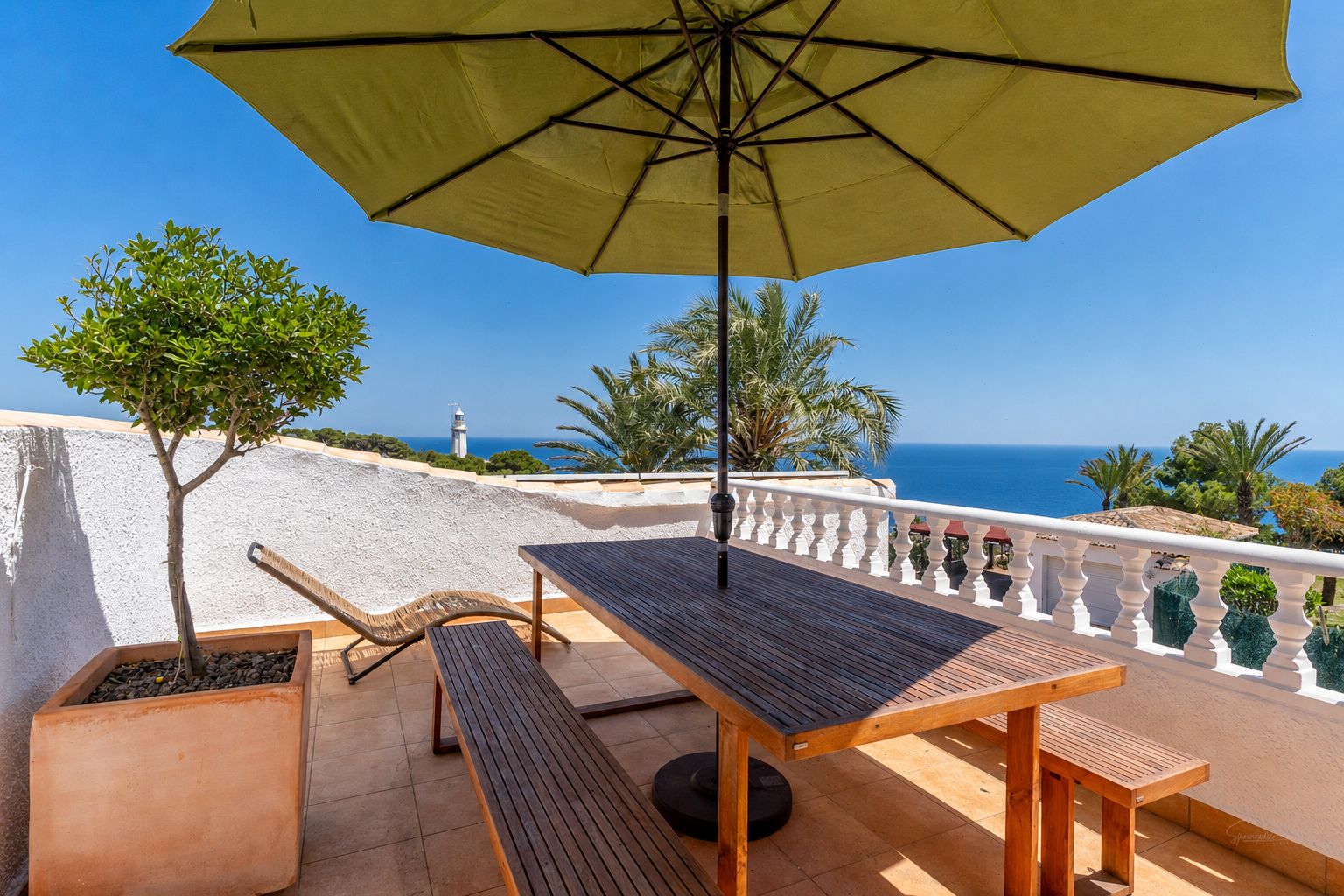 Amplia terraza con mobiliario de comedor de madera y sombrilla, ofreciendo vistas panorámicas al mar y un faro lejano. Suelo de baldosas y árbol en maceta.