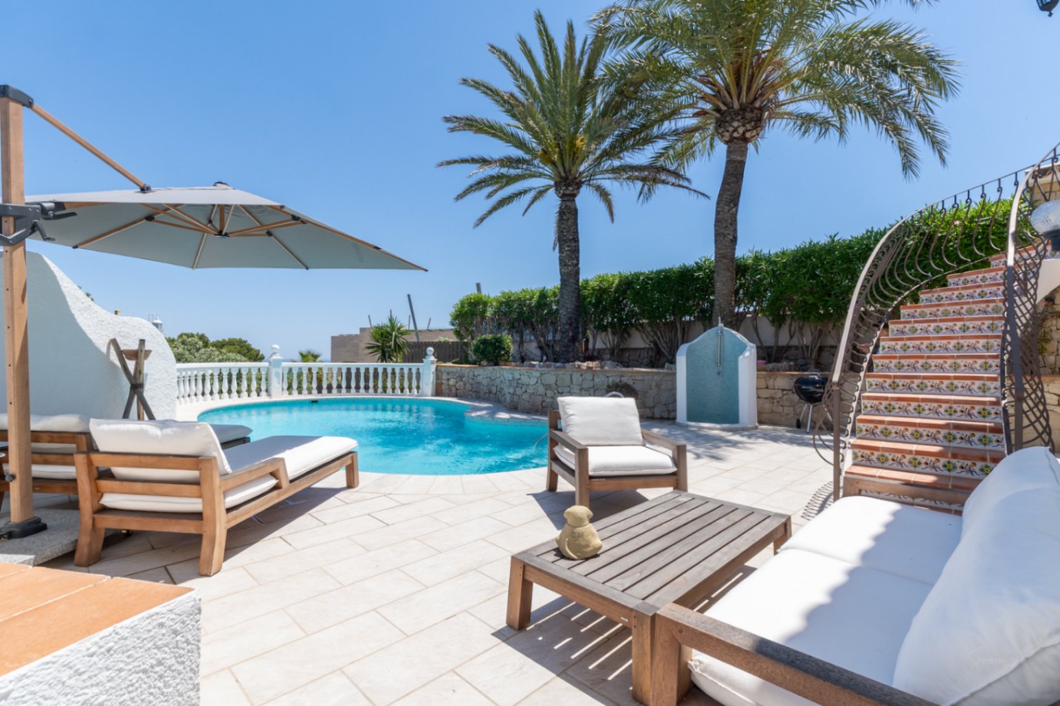 Amplia terraza junto a la piscina con tumbonas de madera, un conjunto de sofás exterior y una escalera curva con azulejos decorativos. Palmeras y una ducha exterior complementan el espacio.