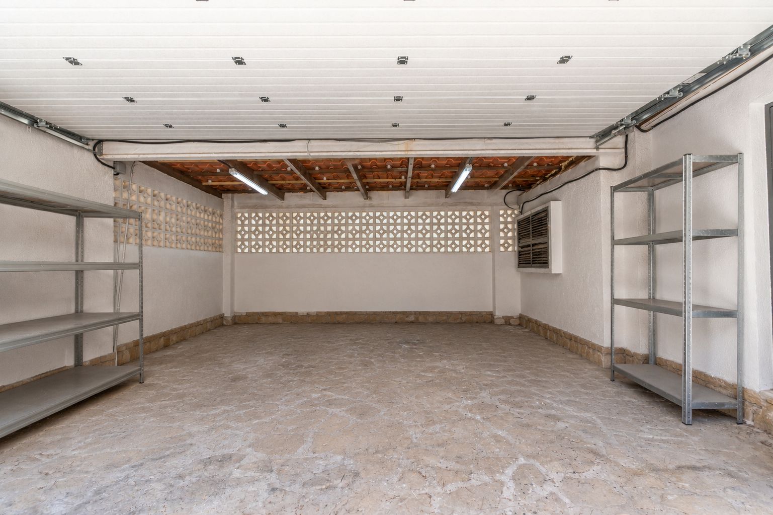 Amplio garaje con suelo de hormigón texturizado, paredes de estuco blanco y bloques decorativos de ventilación. Incluye tejas de terracota vistas, iluminación fluorescente y estanterías metálicas.