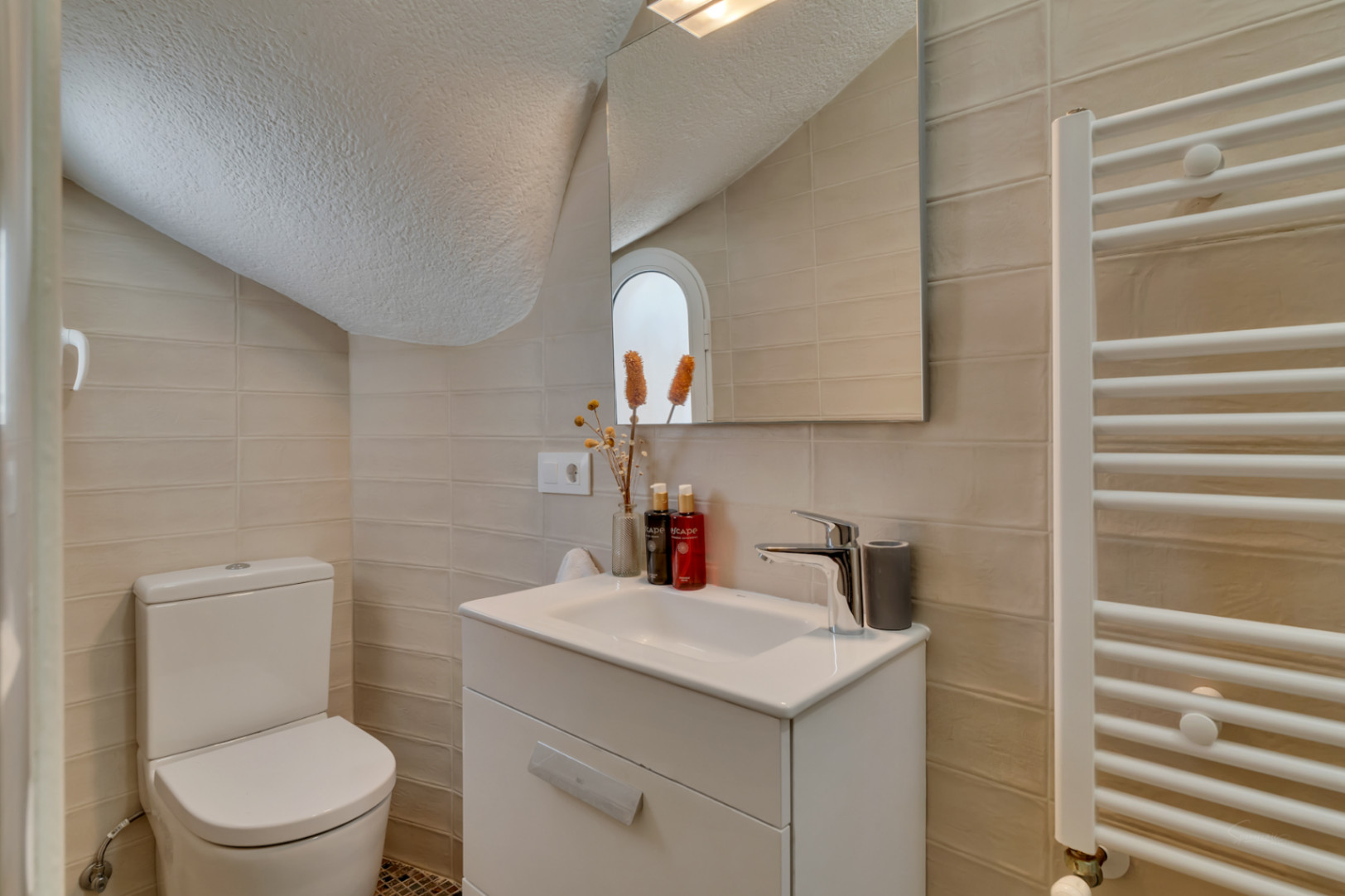 Aseo funcional con paredes de azulejos claros, mueble de lavabo blanco y un inodoro. Un espejo refleja una ventana arqueada y un toallero calefactable.