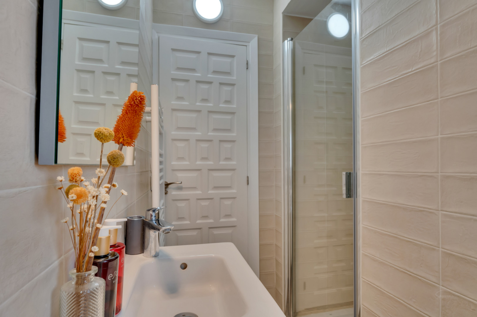 Baño compacto con lavabo blanco, grifería cromada y ducha con mampara de cristal. Destacan los azulejos beige y la puerta panelada.