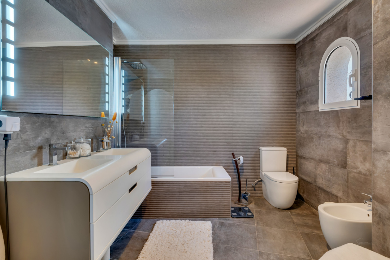 Baño moderno con azulejos grises texturizados, un mueble de doble lavabo flotante y un espejo iluminado. Una ventana arqueada añade carácter sobre la zona del inodoro.