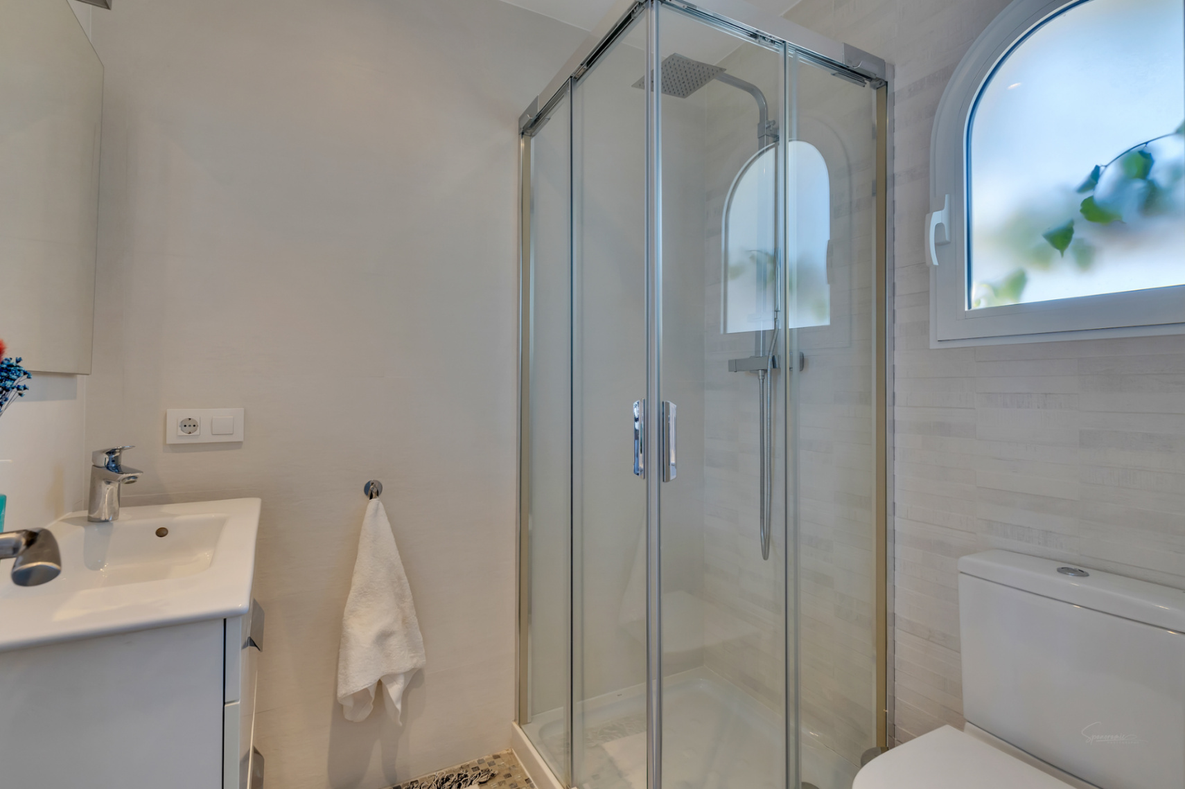 Baño moderno con cabina de ducha de cristal y rociador de lluvia. Destaca una ventana arqueada esmerilada y un lavabo con mueble blanco, junto a azulejos texturizados.