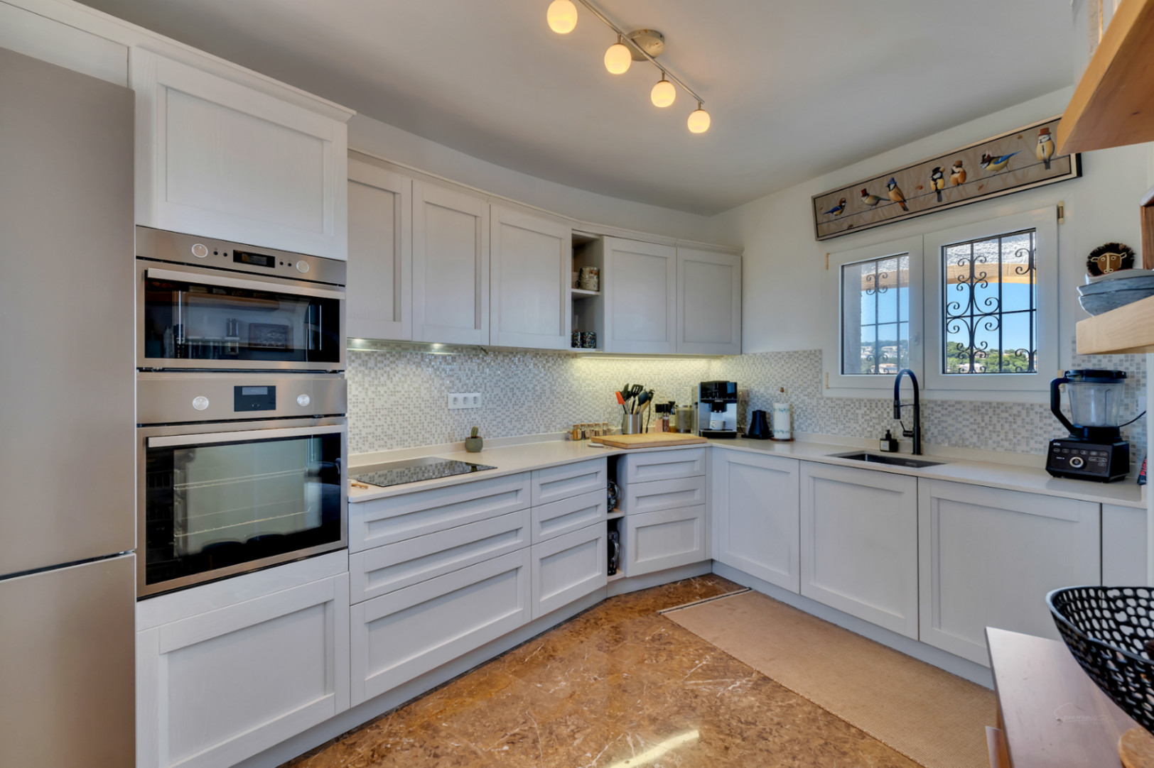 Cocina equipada con armarios blancos estilo shaker, electrodomésticos integrados de acero inoxidable, encimera clara y salpicadero de mosaico. Ventana con rejas decorativas y vistas verdes.
