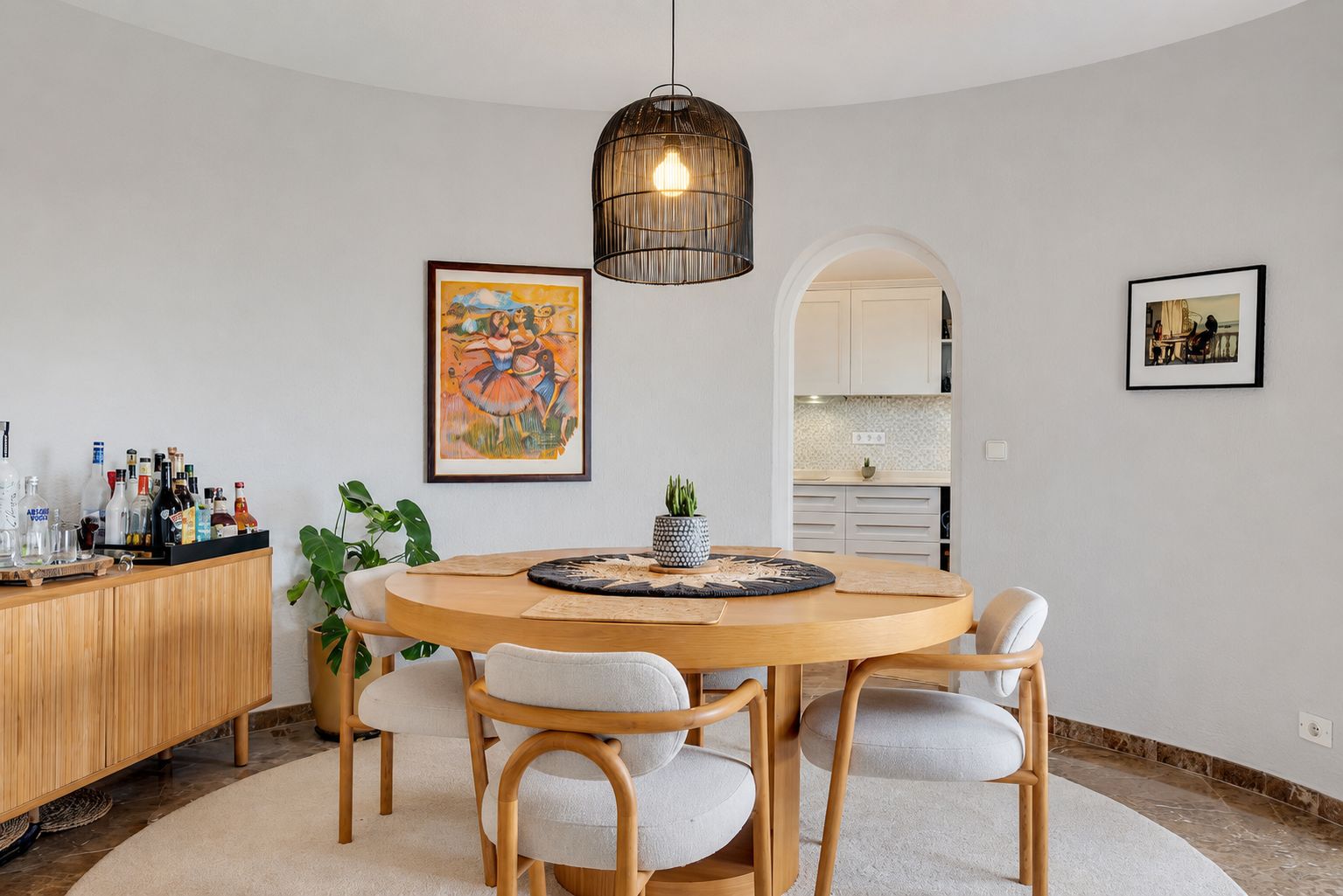 Comedor curvo con mesa redonda de madera y cuatro sillas tapizadas, iluminado por una lámpara colgante de mimbre. Un arco conecta con la cocina.