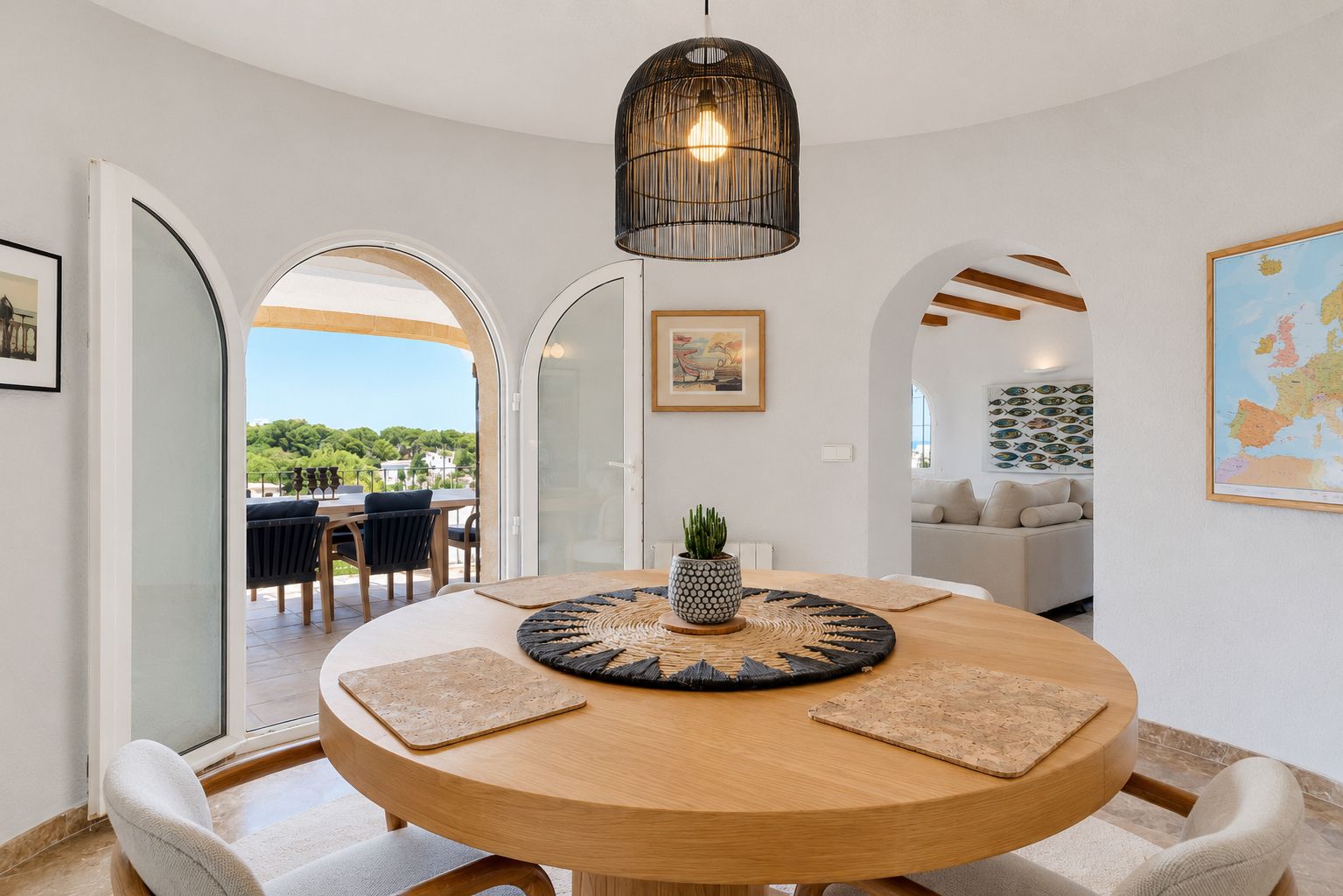 Comedor luminoso con mesa redonda de madera y lámpara de mimbre, conectado a una terraza exterior con vistas a la vegetación. Un arco conduce a la sala de estar.