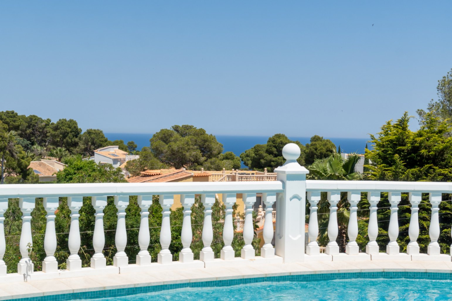 Desde la piscina, una balaustrada blanca enmarca las vistas panorámicas al mar Mediterráneo, tejados de Jávea y exuberante vegetación bajo un cielo azul.